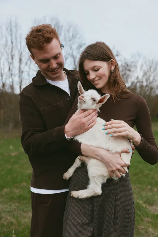 A young couple in casual clothing cradle a white goat kid in their arms in a sunny field, sharing a tender moment. A lifestyle scene showing bond, care, and rural warmth.