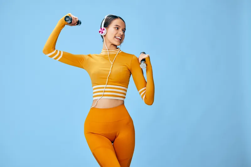 Woman fitness, sport portrait: young woman in bright orange workout set lifting small dumbbells while wearing headphones, smiling confidently in a studio pose against a clean light blue background.