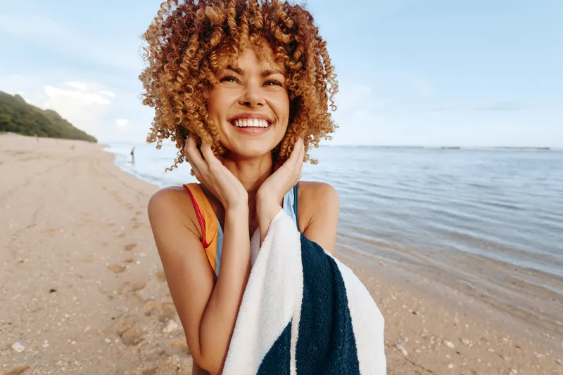 Happy young woman with curly hair smiles brightly on the sandy beach wrapped in a towel with ocean and blue sky in the background during sunny day.