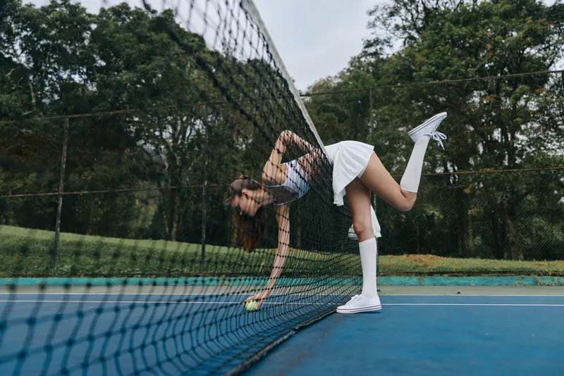 Stylish young woman in a tennis outfit, gracefully reaching for a ball near the net on a sunny day, showcasing active lifestyle and sportsmanship