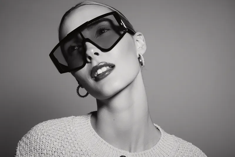 Young Caucasian woman with light skin wearing oversized sunglasses and hoop earrings poses in knitted sweater. Studio portrait in black and white with modern fashion concept.