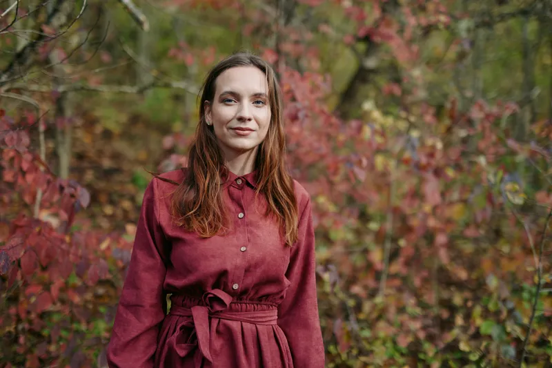 Portrait of a young woman in a simple dress standing among autumn foliage, captured with authentic natural expression and genuine mood, highlighting realistic human presence and quiet calm.