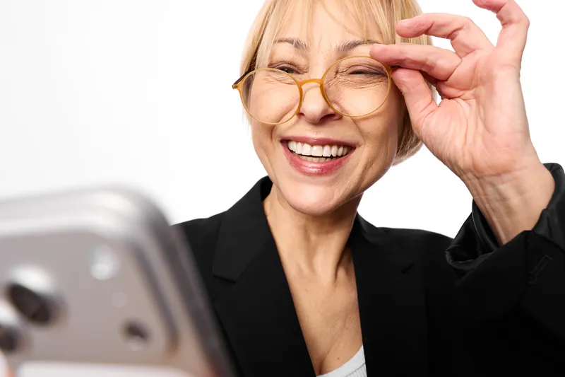 Smiling elderly woman adjusting glasses while looking at a smartphone on desk, close portrait with soft indoor light, technology and communication, joyful mood.