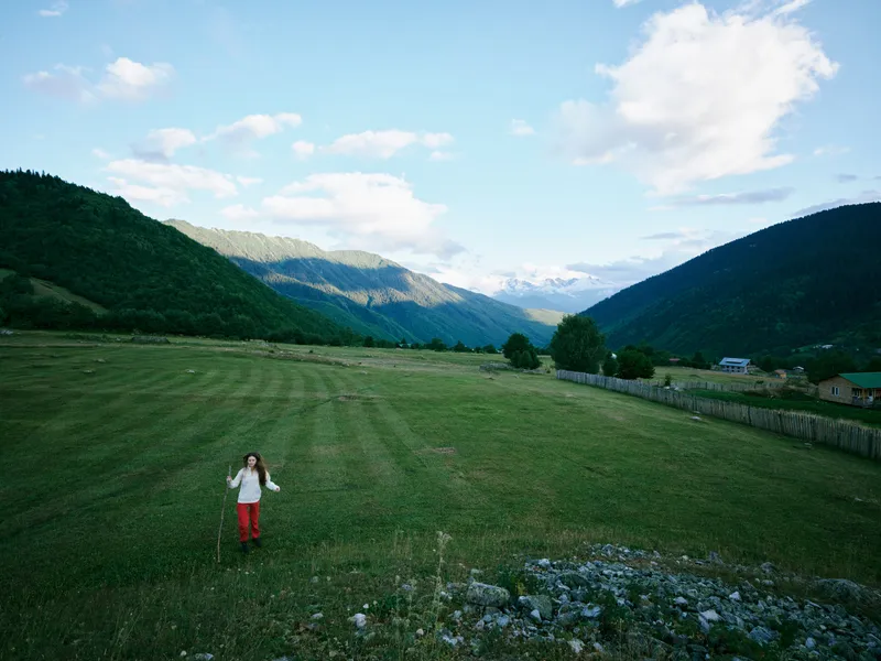 Wide meadow between hills with a girl walking across green grass, distant mountains under a bright sky and clouds, capturing freedom, nature, and outdoor exploration