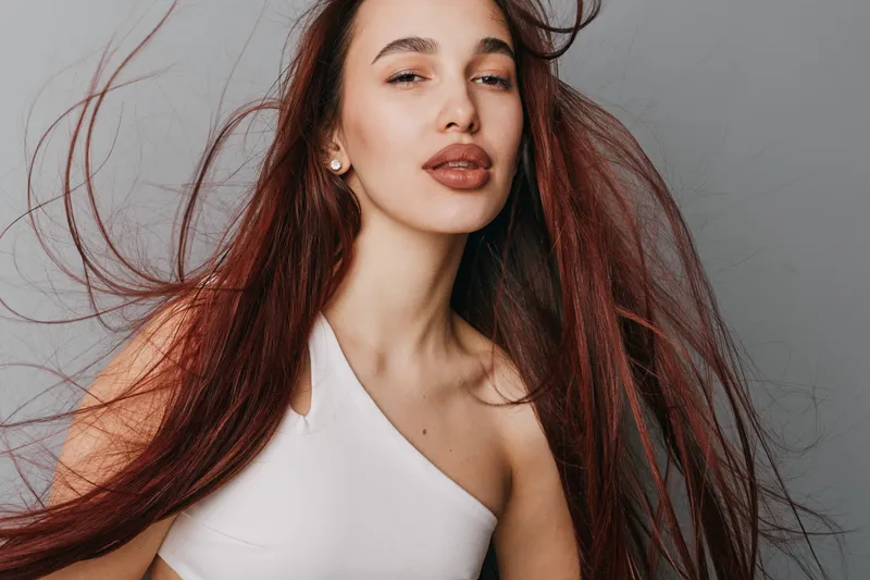 Woman with long hair and natural makeup posing in white top against gray background. Young female model with flowing hair and confident expression in studio portrait.