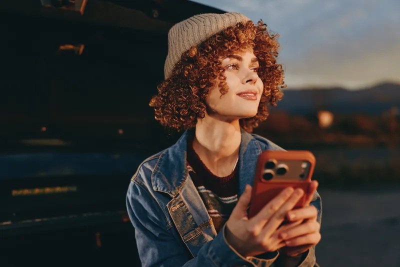 Woman lifestyle portrait outdoors sunset, curly hair, denim jacket, smiling while holding smartphone, casual selfie mood, urban evening night streetlight glow, soft blur behind her