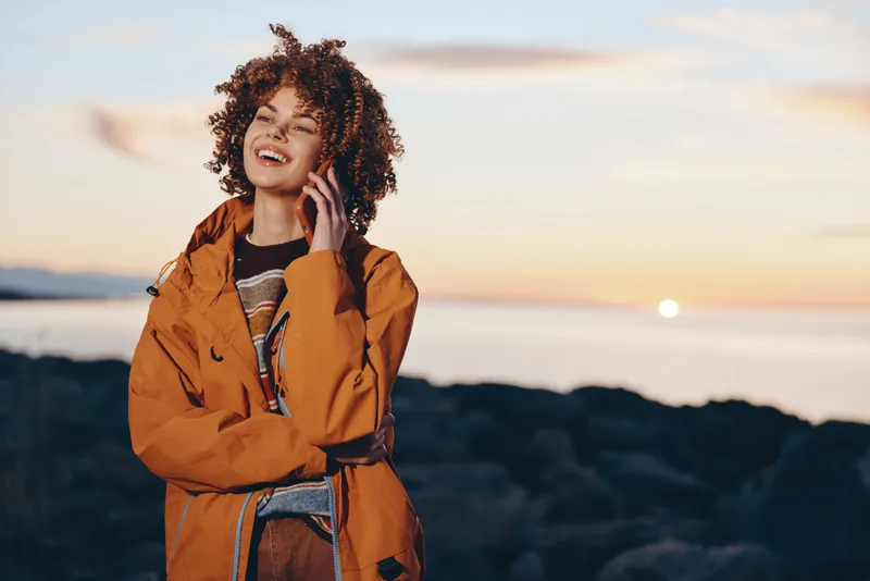 Smiling woman with curly hair wearing an orange jacket outdoors at sunset on mountains viewpoint, lifestyle mood with warm light and nature scenery for travel and relaxation