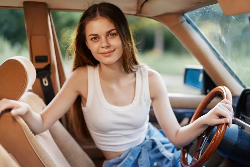 Young woman smiling in a vintage car, wearing a casual white tank top and denim shorts, embodying a carefree summer vibe and a sense of adventure.