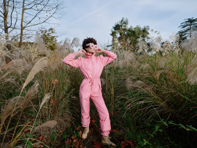 Female in pink jumpsuit posing in tall grasses outdoors, capturing playful energy amid open field and natural scenery under a clear sky with wind in hair