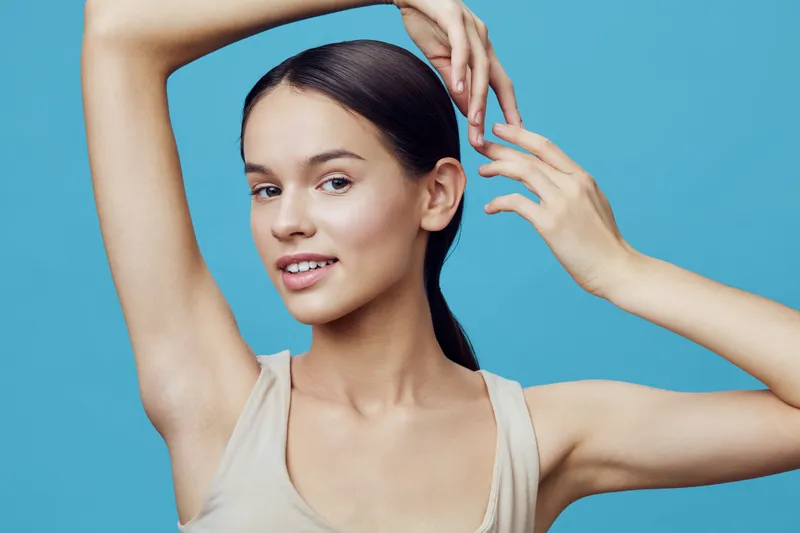 Young model with long hair poses confidently against a blue background, showcasing beauty and elegance while applying skincare products for a glowing complexion