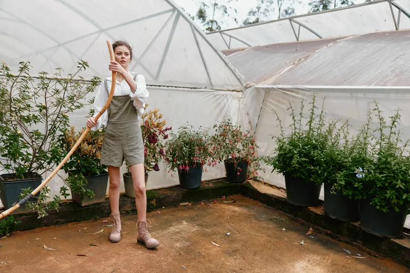 Young woman in a green house wearing a stylish jumpsuit, holding a gardening tool, showcasing sustainable living and eco friendly gardening concepts