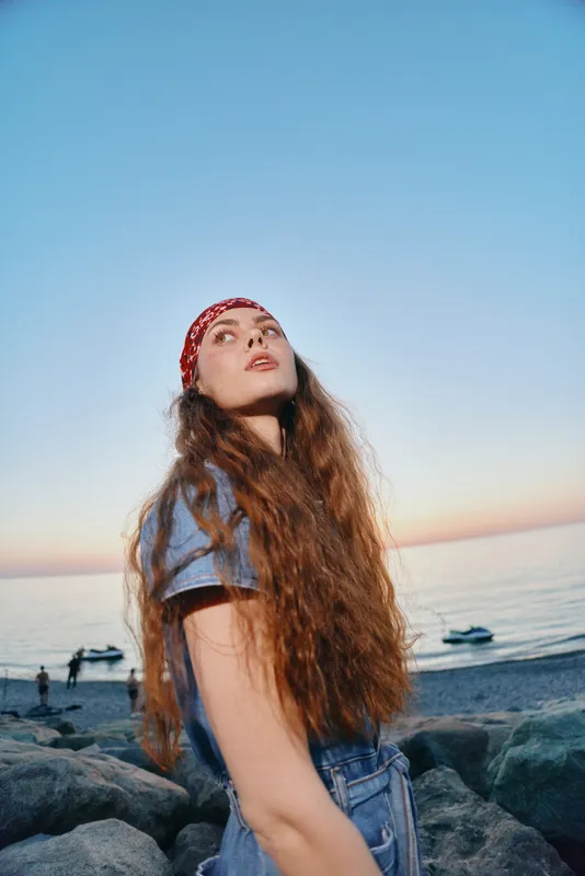 Lifestyle woman in modern boho-western denim street style with long hair and red bandana by the ocean at sunset, film color tones creating a warm and relaxed vibe.
