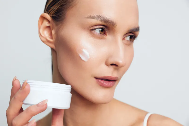 A young woman with clear skin applies white cream on her cheek holding an open jar. Close-up of facial skincare routine for moisturizing and healthy skin care concept.