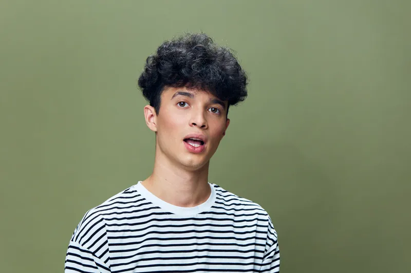Young man with curly hair wearing a striped shirt against a muted green background, expressing surprise or curiosity in casual portrait style.