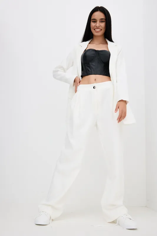 smiling woman in white pants and jacket with black crop top posing confidently against white background, casual style, long hair, indoor lighting