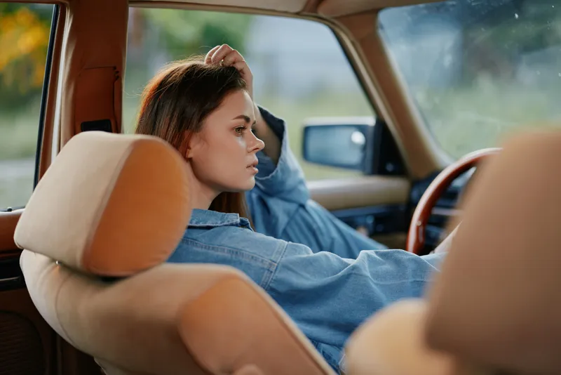 Young woman in denim shirt relaxed in vintage car interior, capturing a moment of contemplation and serenity in nature, featuring warm tones and soft lighting.