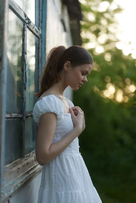 Young woman in a white dress standing by a window, reflecting on life in a soft green background, evoking calm and tranquility during golden hour
