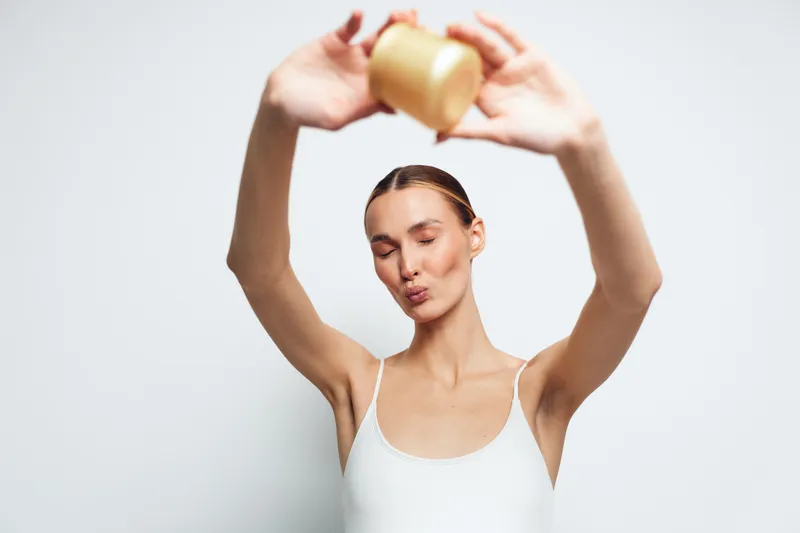 Young woman in a white tank top holds a jar of cream above her head while making a playful face against a plain white background.