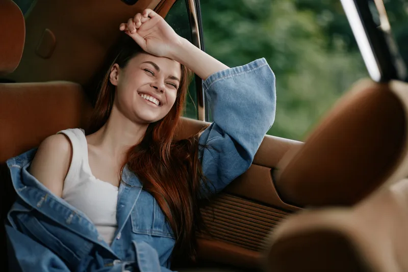 Young woman with long red hair laughing joyfully in a vintage car, wearing a denim jacket and a white tank top, capturing a carefree summer vibe.