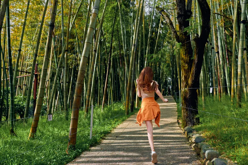 Woman running along a bamboo forest path in an orange dress, back view on a gravel trail surrounded by green trees and sunlight, motion and summer outdoor adventure.