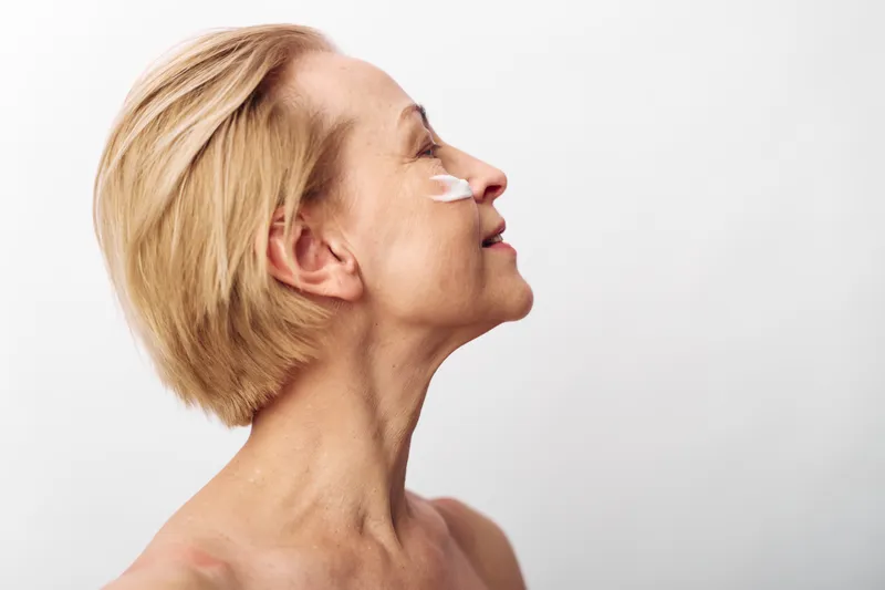 Mature woman with short blonde hair applies white facial cream on cheek, smiling softly. Concept of skincare, aging skin, beauty routine, self-care, and natural health care.