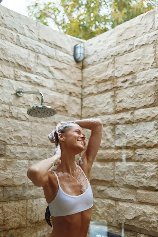 smiling woman enjoying outdoor shower with water falling, surrounded by textured stone wall and natural light, fresh and relaxed summer moment