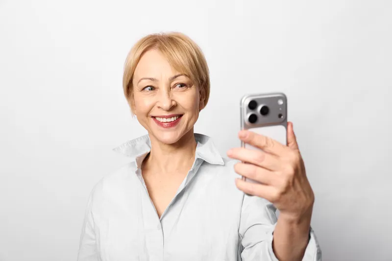 Woman smiling smartphone selfie portrait communication technology friendly studio white background, mature blond woman in blouse holding mobile, closeup casual indoor shot, digital communication