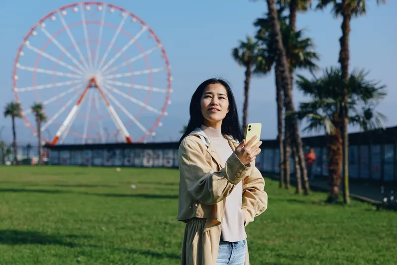 Woman holding smartphone in a sunny park with a ferris wheel behind, casual jacket, green grass, palm trees, relaxed leisure moment, outdoor city vibe, candid street style