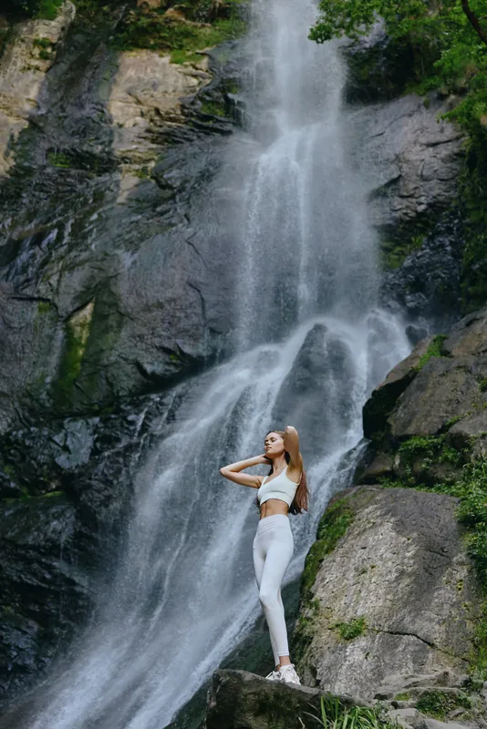 athletic woman in white sportswear standing by a large waterfall in a lush green forest, showcasing strength and nature connection during outdoor fitness.
