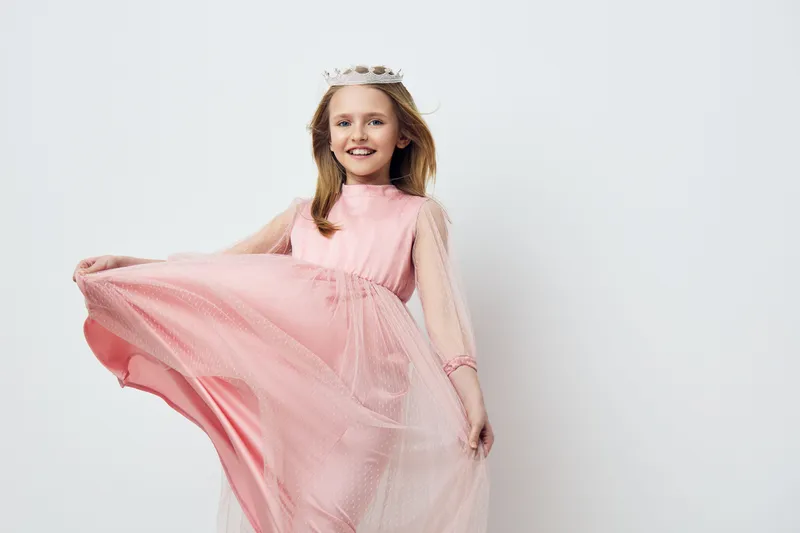 A joyful young girl wearing a pink dress and a crown, smiling and playfully twirling, ideal for concepts of childhood, fashion, and celebration