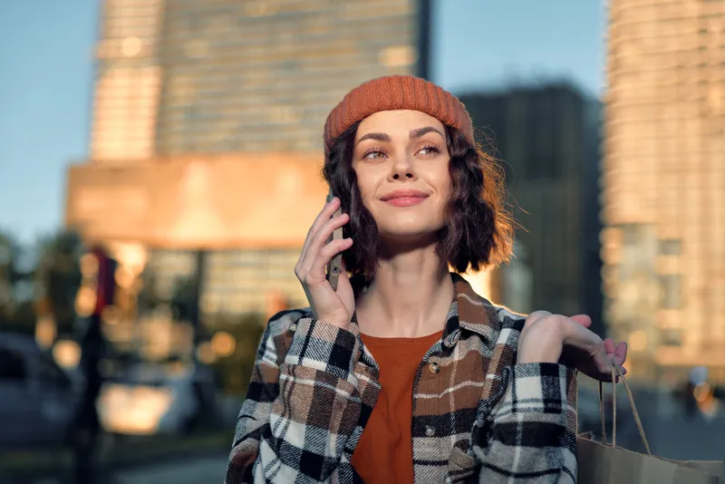 Woman shopping in city, phone to ear and portrait smile with paper bag, urban golden hour glow and authenticity, candid lifestyle moment reflecting mindful living and emotional storytelling.