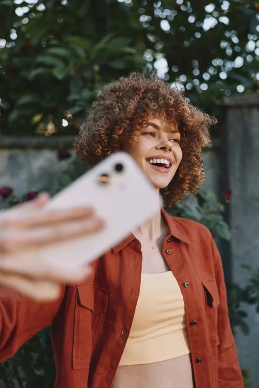 Young woman with curly hair smiles while taking a selfie outdoors, wearing a burnt orange jacket over a pale yellow crop top and standing in natural light