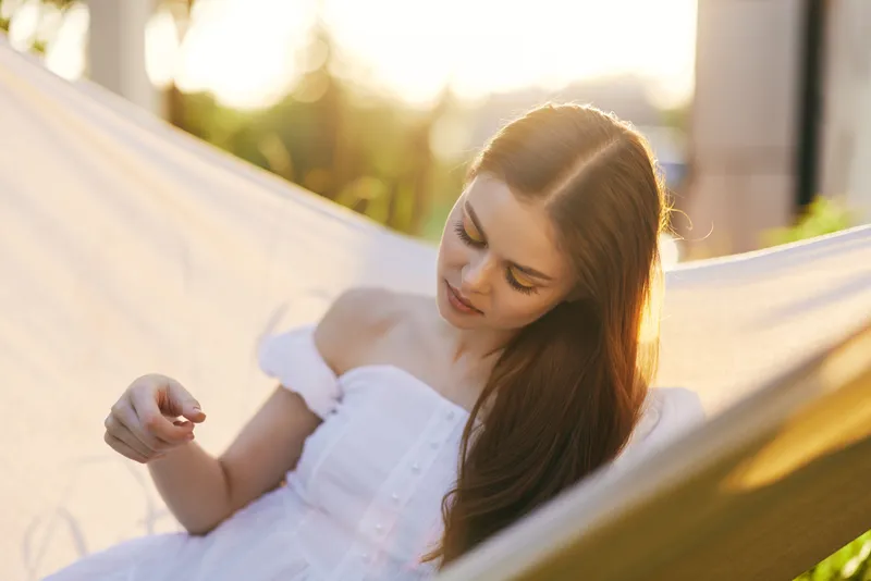A young woman relaxing in a hammock during sunset, wearing a white dress, exuding a peaceful aura, surrounded by nature Ideal for concepts of relaxation and tranquility