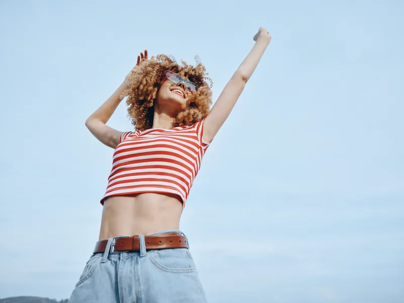Young person dancing outdoors in a red and white striped crop top with light denim jeans, raised arms, joyful motion, clear blue sky, carefree summer vibe.
