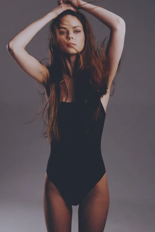 Young woman in black bodysuit posing with hands raised, long hair flowing and serious expression, minimalistic studio background.
