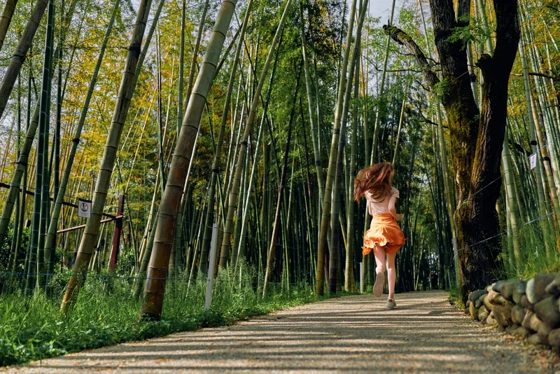 Woman running along a winding path through a dense bamboo forest, back view in a flowing orange dress. Motion blur, sunlight dappled trail, summer nature scene and carefree escape.