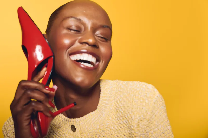 Happy African woman with dark skin laughs holding a red high heel shoe near her face. Bright yellow background and casual beige sweater create a cheerful, playful mood for fashion concept.