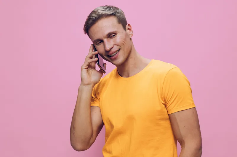 Smiling young man in yellow t-shirt talking on mobile phone, casual lifestyle, happy expression, indoor setting, pink background, positive mood, relaxed atmosphere, modern style.