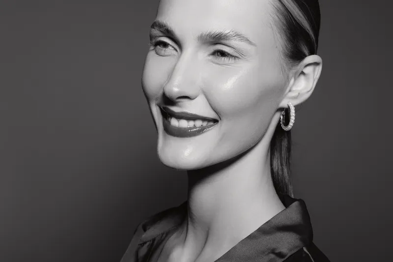 Smiling young Caucasian woman with fair skin and slicked back hair wears hoop earrings and dark satin blouse. Studio portrait in black and white with soft lighting and confident mood.