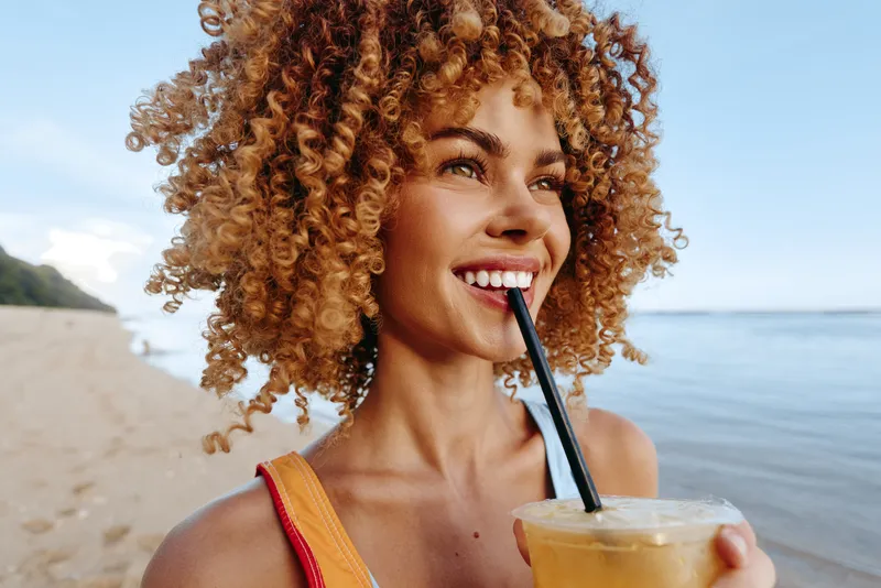 Young woman with curly hair enjoys a cold drink on a sunny beach while smiling and looking into the distance with a relaxed and happy expression.