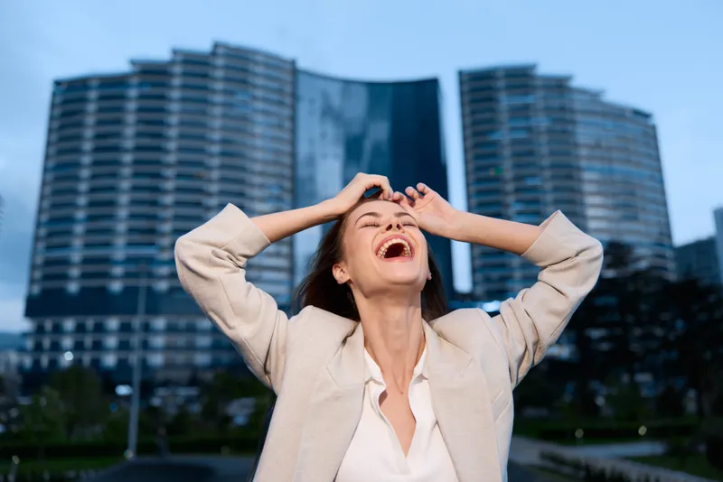 joyful woman laughing outdoors in beige blazer with modern city building background expressing happiness and freedom in urban business style