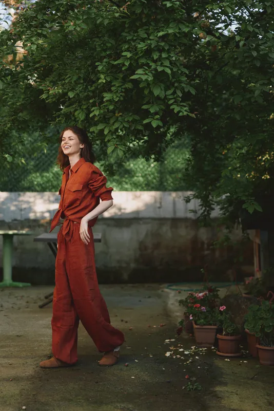 Smiling woman in a rust jumpsuit strolling through a sunny garden, showcasing relaxed lifestyle and outdoor fashion. Warm natural light highlights casual confidence and candid charm