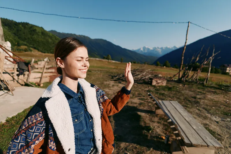 Girl smile countryside mountains sunlight jacket outdoors portrait, young woman standing with eyes closed and hand raised, enjoying warm sun and rustic rural landscape on clear day
