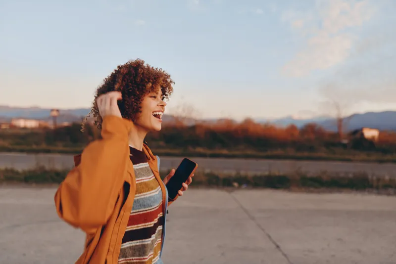 Woman lifestyle outdoors at sunset, smiling with curly hair, orange jacket, holding a phone, city background with warm sunlight, casual street moment, relaxed joyful mood