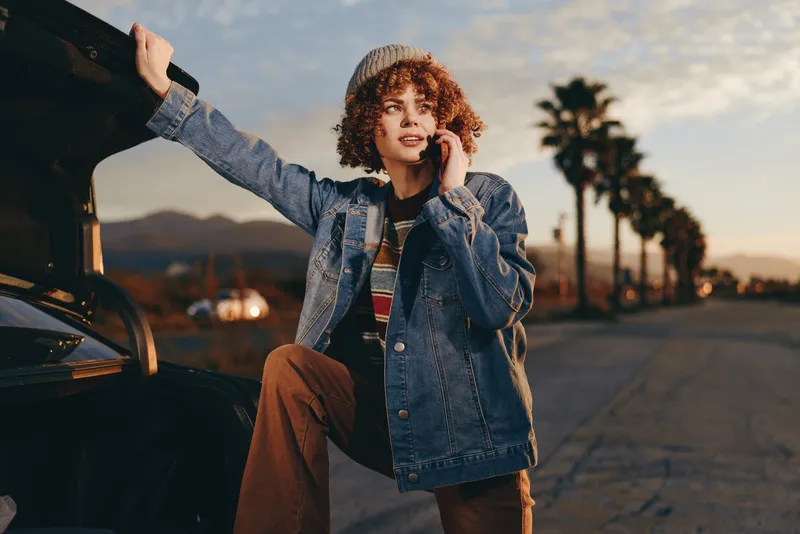 Woman with curly hair, wearing a denim jacket, standing beside a car at sunset, one hand on the open door, outdoors lifestyle for travel, warm evening light