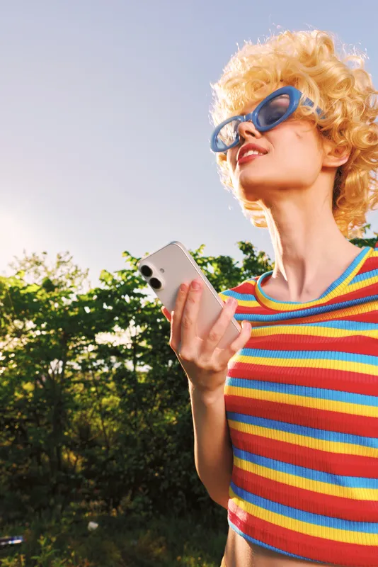 Smiling woman with curly blonde hair wearing a colorful striped shirt outdoors, holding a smartphone. Vintage film color mood, bright sun, and carefree lifestyle in a playful summer scene.