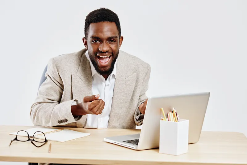 Professional young African American man in beige blazer smiling cheerfully working on laptop at desk in bright office environment with eyeglasses and stationery. People business success concept