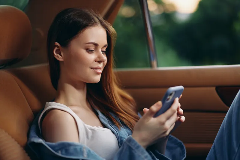 Young woman smiling while using smartphone in car, casual outfit with denim jacket, relaxed expression, natural background, vibrant colors, lifestyle concept.