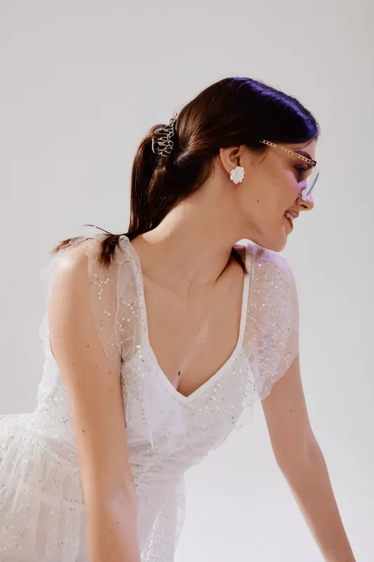 Elegant woman in a white lace dress and stylish glasses smiles gently while leaning forward. Studio shot on a white background. Concept of beauty, fashion, and elegance.