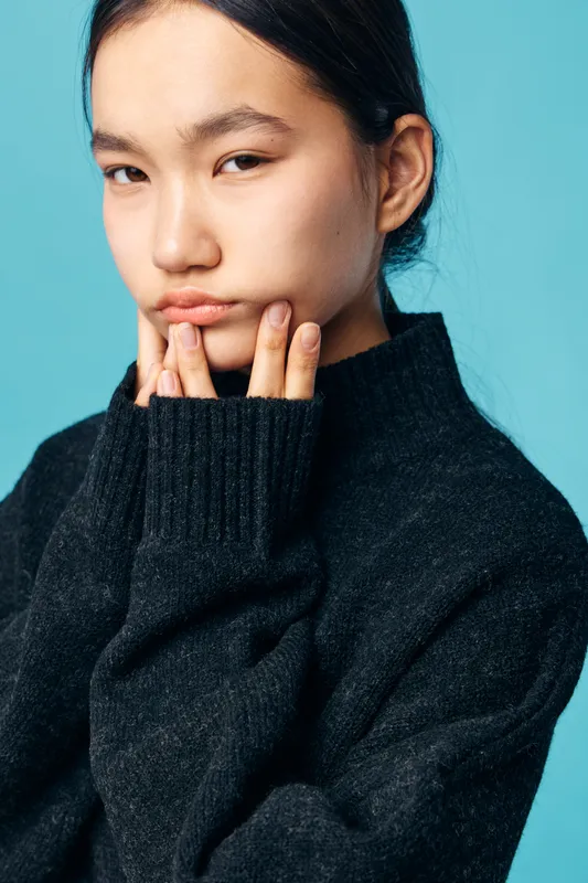 Portrait of young woman in black sweater with fingers touching chin looking thoughtful and serious, studio shot with blue background for fashion or emotion concept.